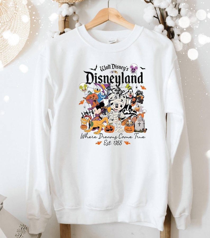 May include: White crew neck sweatshirt with a Halloween-themed Disney graphic. The design features various Disney characters dressed in costumes, including Mickey Mouse and Donald Duck. The text reads "Walt Disney's Disneyland" and "Where Dreams Come True Est. 1955".