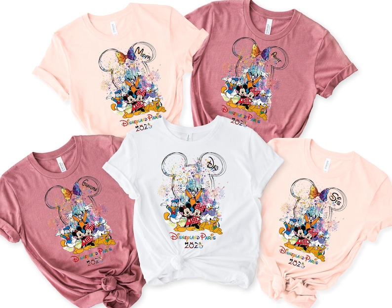 May include: Four t-shirts with a Mickey Mouse and friends design. The shirts are pink, white, and a darker pink. The design features Mickey Mouse, Minnie Mouse, Donald Duck, Goofy, and Pluto in front of the Sleeping Beauty Castle with fireworks. The text on the shirts says "Disneyland Paris 2023" and includes the words "Mom", "Dad", "Sister", and "Aunt".