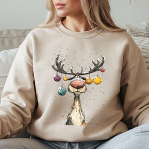 May include: A beige crewneck sweatshirt featuring a whimsical illustration of a reindeer with a large pink nose. The reindeer has colourful Christmas ornaments hanging from its antlers, and small white dots representing falling snow.