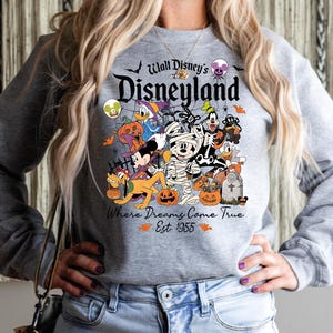 May include: Grey sweatshirt with a Halloween Disney design. It features Mickey Mouse, Donald Duck, and Goofy, alongside pumpkins and bats. The text reads "Walt Disney's Disneyland" and "Where Dreams Come True Est. 1955" on the front.