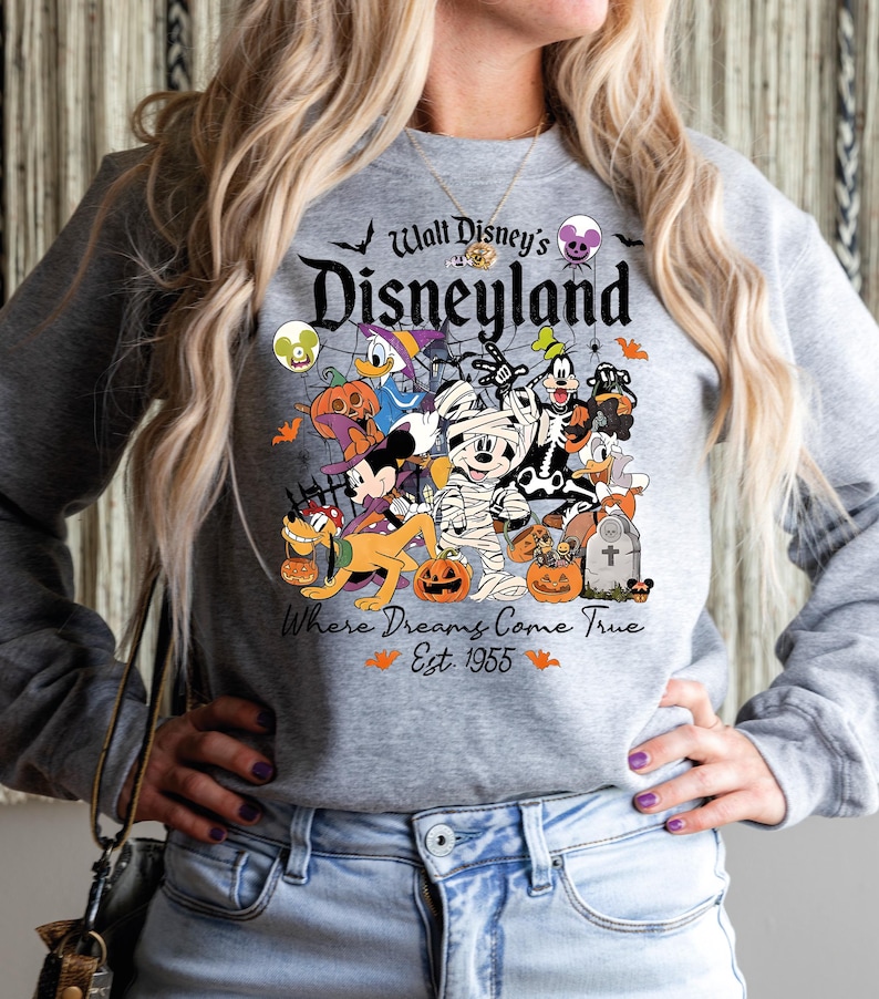 May include: Grey sweatshirt with a Halloween Disney design. It features Mickey Mouse, Donald Duck, and Goofy, alongside pumpkins and bats. The text reads "Walt Disney's Disneyland" and "Where Dreams Come True Est. 1955" on the front.