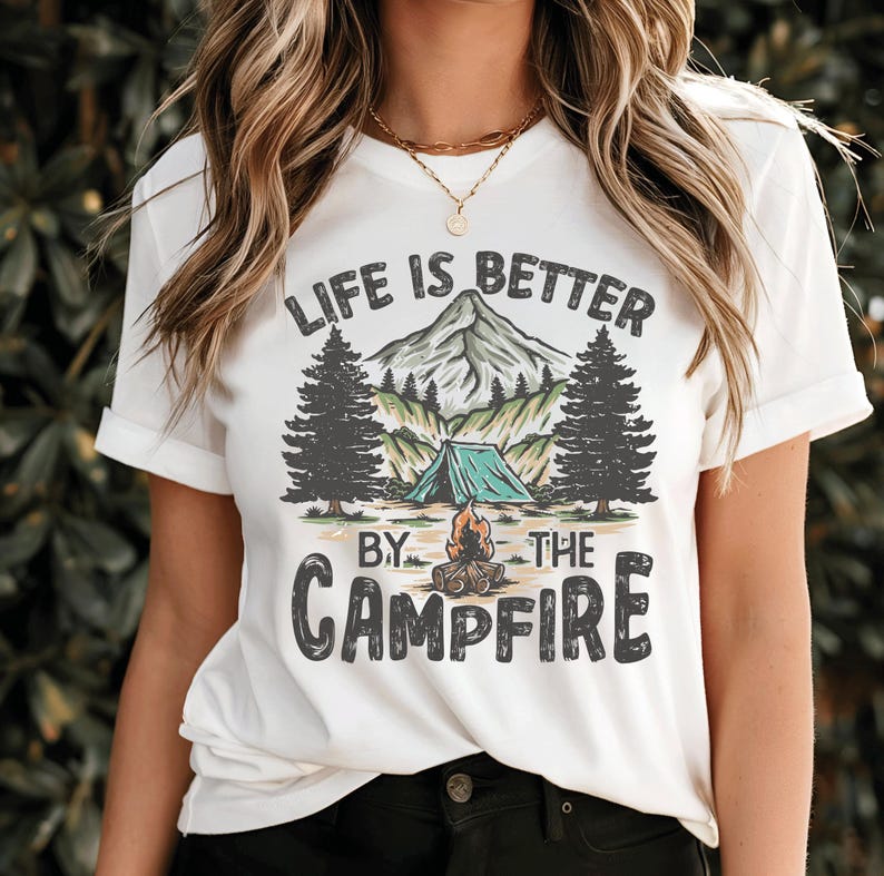 May include: White t-shirt with a camping graphic. The graphic features a mountain, trees, a tent, and a campfire, with the text "LIFE IS BETTER BY THE CAMPFIRE" in a stylized font. The shirt has short sleeves.