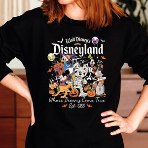 May include: Black long-sleeved sweatshirt with a colourful Halloween graphic of Disney characters. The graphic includes Mickey Mouse as a mummy, Donald Duck, and other characters with pumpkins and bats. Text reads "Walt Disney's Disneyland" and "Where Dreams Come True Est. 1955."