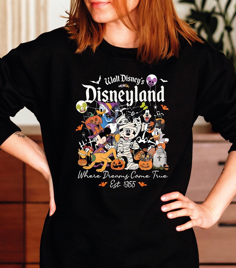 May include: Black long-sleeved sweatshirt with a colourful Halloween graphic of Disney characters. The graphic includes Mickey Mouse as a mummy, Donald Duck, and other characters with pumpkins and bats. Text reads "Walt Disney's Disneyland" and "Where Dreams Come True Est. 1955."