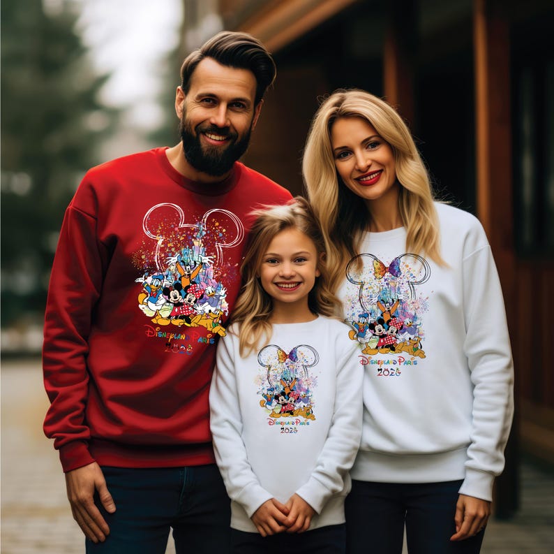 May include: A family in matching sweatshirts featuring a Disney design. The red sweatshirt has a Mickey Mouse design with characters and the text "Disneyland Paris 2026". The white sweatshirts have a similar design.