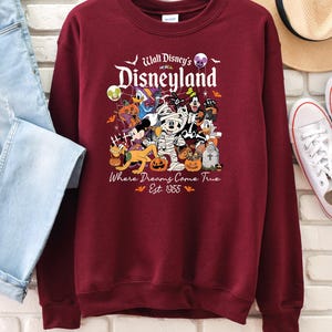 May include: Burgundy crew neck sweatshirt with a Disneyland Halloween design. The graphic features Mickey Mouse, Donald Duck, and other Disney characters in Halloween costumes. Text reads "Walt Disney's Disneyland" and "Where Dreams Come True Est. 1955".