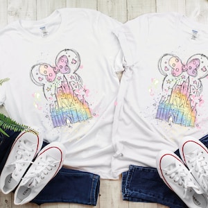 May include: Two white t-shirts with a colorful sketch of Cinderella's castle and Minnie Mouse ears on them. The shirts are laid out on a wooden surface with a pair of white sneakers on each side.