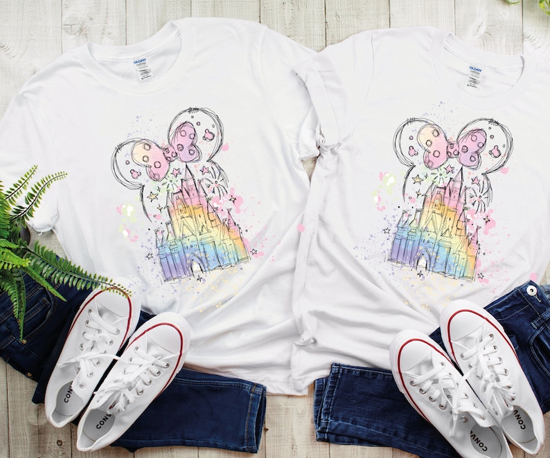 May include: Two white t-shirts with a colorful sketch of Cinderella's castle and Minnie Mouse ears on them. The shirts are laid out on a wooden surface with a pair of white sneakers on each side.