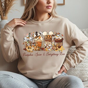 May include: Beige sweatshirt featuring a cartoon design of pumpkin spice drinks and characters. The text "Pumpkin Spice & Everything Nice" is printed in a cursive font. The design includes pumpkins and autumn leaves.