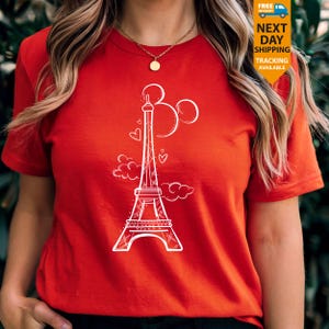 K&ouml;nnte beinhalten: Ein rotes T-Shirt mit einer wei&szlig;en Grafik des Eiffelturms, Mickey Mouse Ohren und Herzen. Das Design ist eine skurrile Mischung aus Pariser und Disney-Themen. Das Shirt hat kurze &Auml;rmel und einen Rundhalsausschnitt.