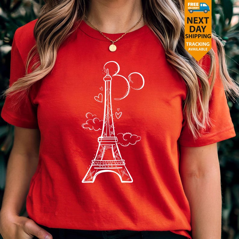 K&ouml;nnte beinhalten: Ein rotes T-Shirt mit einer wei&szlig;en Grafik des Eiffelturms, Mickey Mouse Ohren und Herzen. Das Design ist eine skurrile Mischung aus Pariser und Disney-Themen. Das Shirt hat kurze &Auml;rmel und einen Rundhalsausschnitt.