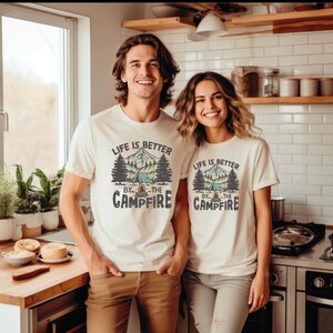 May include: Two people wearing off-white t-shirts with the text "LIFE IS BETTER BY THE CAMPFIRE" printed on the front. The graphic includes a mountain scene, trees, and a tent. The people are standing in a kitchen.