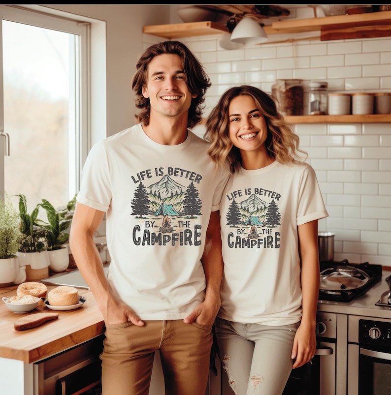 May include: Two people wearing off-white t-shirts with the text "LIFE IS BETTER BY THE CAMPFIRE" printed on the front. The graphic includes a mountain scene, trees, and a tent. The people are standing in a kitchen.