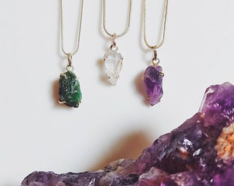 Puede incluir: Tres collares de plata con diferentes colgantes de piedras preciosas en bruto. Los colgantes son una esmeralda verde, una amatista morada y un cristal de cuarzo transparente. Los collares se muestran sobre un fondo de cristales de amatista morados.