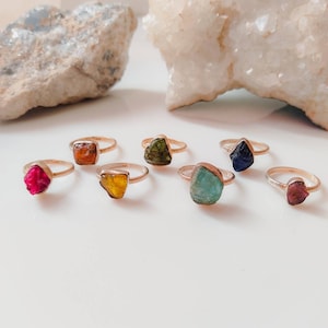 Könnte beinhalten: Sieben goldene Ringe mit verschiedenen farbigen Edelsteinen. Die Ringe haben verschiedene Formen und Größen von Edelsteinen, darunter ein Quadrat, ein Dreieck und ein Oval. Die Ringe sind in einer Reihe auf einer weißen Oberfläche angeordnet.