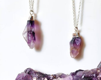 May include: Two silver necklaces with amethyst crystal pendants. The crystals are raw and have a purple and white color. The necklaces are displayed on a bed of amethyst crystals.