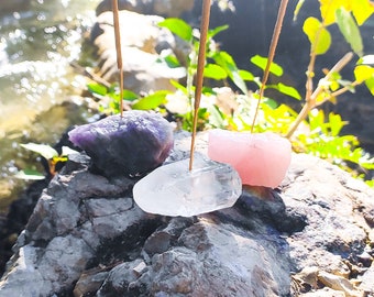 May include: Three incense sticks are burning on top of three different colored crystals. The crystals are purple, pink, and clear. The crystals are sitting on a gray rock.