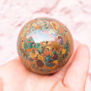 Puede incluir: Una esfera redonda, pulida, de color marrón con motas verdes, amarillas y rojas. La esfera está hecha de piedra natural y tiene una superficie lisa y brillante.