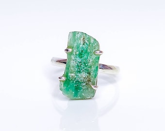May include: A silver ring with a large, raw emerald gemstone. The emerald is a deep green color and has a rough, natural texture.