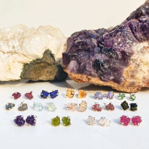 Peut inclure: Assortiment de boucles d'oreilles en pierres précieuses brutes de différentes couleurs, notamment bleu, vert, rouge, violet et orange. Les boucles d'oreilles sont présentées sur une surface blanche avec deux gros rochers en arrière-plan.