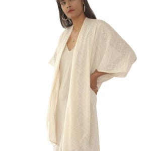 Könnte beinhalten: Eine cremeweiße, vorne offene Strickjacke im Kimono-Stil aus leichtem, strukturiertem Stoff mit feinem Webmuster. Das Kleidungsstück hat weite Dreiviertelärmel und eine lockere, fließende Silhouette, die über einem passenden ärmellosen Kleid liegt.
