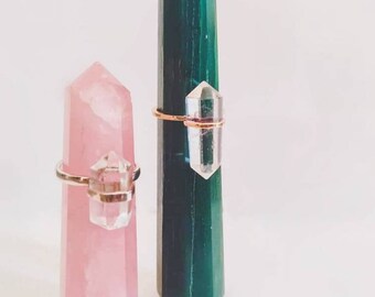 May include: Two crystal point rings on display on crystal point stands. The stands are pink and green.