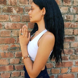 May include: A woman with long black hair is wearing a white tank top and a blue and gold patterned skirt. She is wearing a bracelet with multiple stones on her left wrist and a ring on her right hand. She is standing in front of a brick wall.
