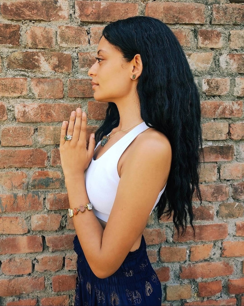 May include: A woman with long black hair is wearing a white tank top and a blue and gold patterned skirt. She is wearing a bracelet with multiple stones on her left wrist and a ring on her right hand. She is standing in front of a brick wall.