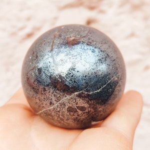 Könnte beinhalten: Eine glatte, dunkelgraue Hämatitkugel mit einem metallischen Glanz und subtilen silbernen Streifen. Die Kugel ist etwa so groß wie ein Golfball.