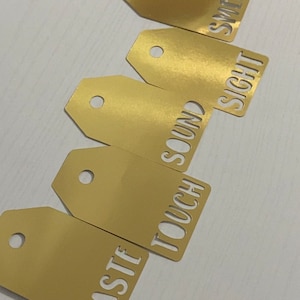 May include: Five gold-colored tags with the words "Taste", "Touch", "Sound", "Sight", and "Smell" cut out of them.