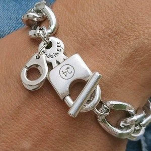 Puede incluir: Pulsera de cadena de plata con un dije de candado, un dije de llave y un dije de esposas. El candado tiene las letras "J.C." grabadas. El dije de llave tiene el texto "made in EEC" grabado.