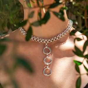 May include: A woman wearing a black tank top with a silver chain necklace featuring three connected circles. The necklace is visible through the green leaves of a tree.