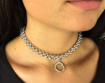 May include: A silver choker necklace with a large, round, silver pendant. The necklace is made of small, linked, silver balls.
