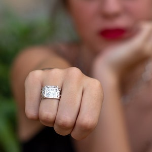 Può includere: Un anello d'argento con una faccia rettangolare, con un design dettagliato. L'anello è indossato su una mano, con lo sfondo sfocato. L'anello sembra essere un pezzo importante.