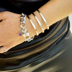 Può includere: Un braccialetto con ciondoli in argento con un ciondolo a forma di lucchetto e una pietra preziosa viola. Il braccialetto è indossato su un polso con tre braccialetti in argento.