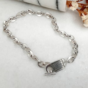 Può includere: Un braccialetto a catena argentata con un design a maglie ovali. Il braccialetto presenta un'etichetta rettangolare con testo inciso e una chiusura a levetta. Il braccialetto è esposto su una superficie bianca con fiori secchi e una pietra scura.