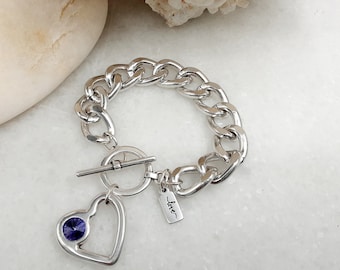 Può includere: Un braccialetto a catena d'argento con un ciondolo a forma di cuore e un cristallo viola. Il ciondolo ha anche la parola "love" incisa.