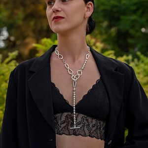 May include: A woman wearing a black blazer, a black lace bralette, and blue jeans. She is wearing a silver chain necklace with a pendant.