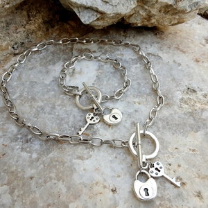 May include: A silver chain necklace with a silver padlock charm and a silver key charm. The necklace has a toggle clasp and a silver bracelet with a padlock charm.