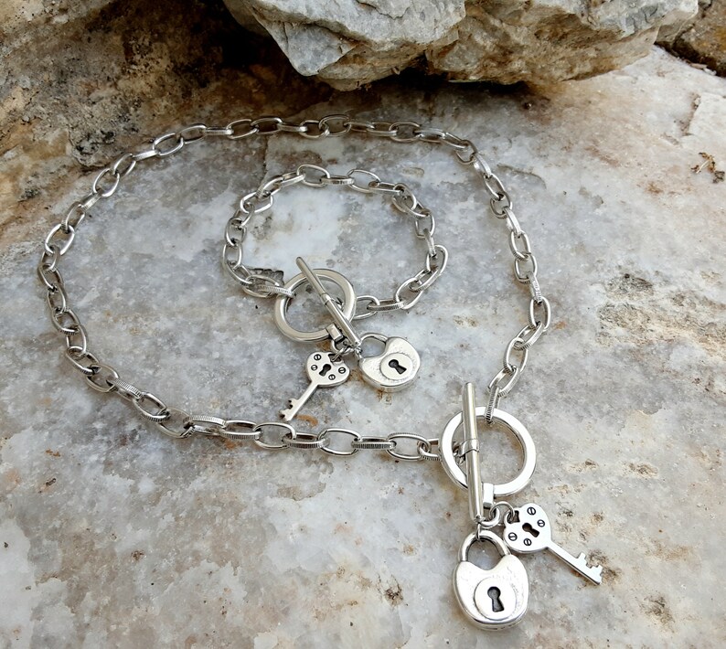 May include: A silver chain necklace with a silver padlock charm and a silver key charm. The necklace has a toggle clasp and a silver bracelet with a padlock charm.