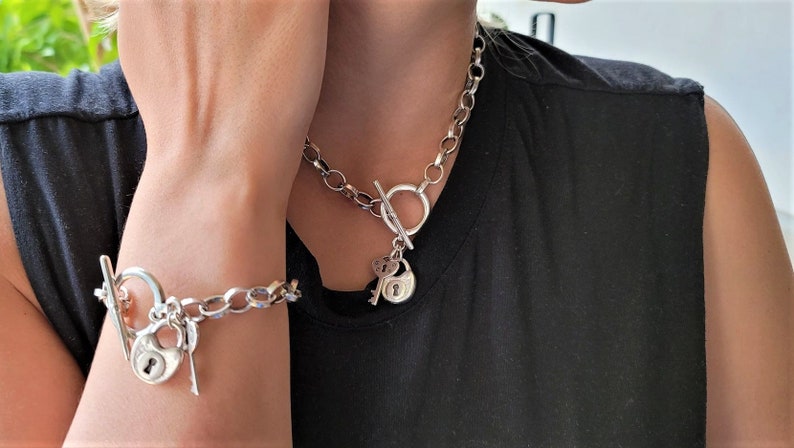 May include: A silver chain necklace and bracelet with a padlock and key charm. The necklace and bracelet have a toggle clasp.