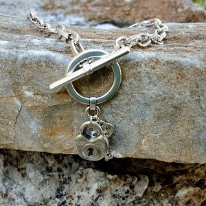 May include: A silver necklace with a toggle clasp and a charm featuring a padlock and keys. The necklace is on a rock background.