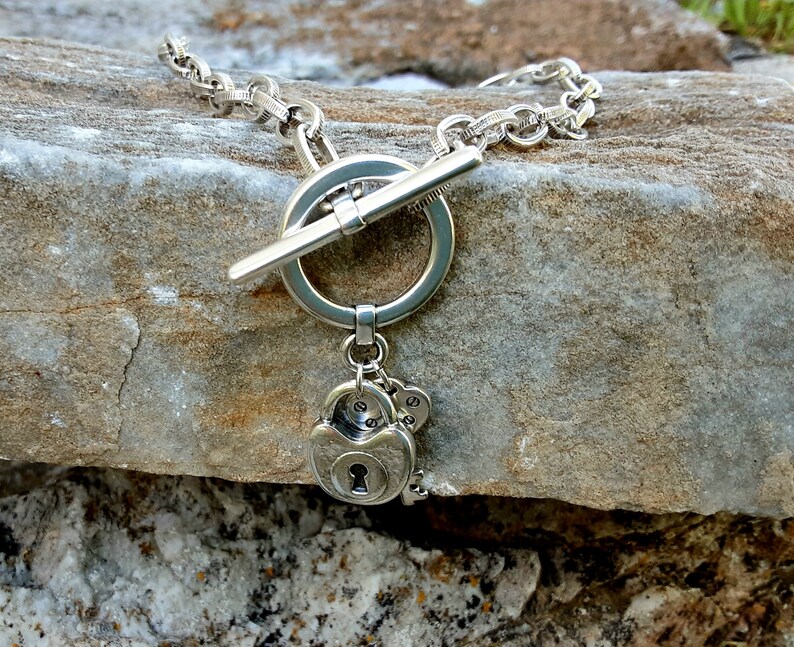 May include: A silver necklace with a toggle clasp and a charm featuring a padlock and keys. The necklace is on a rock background.