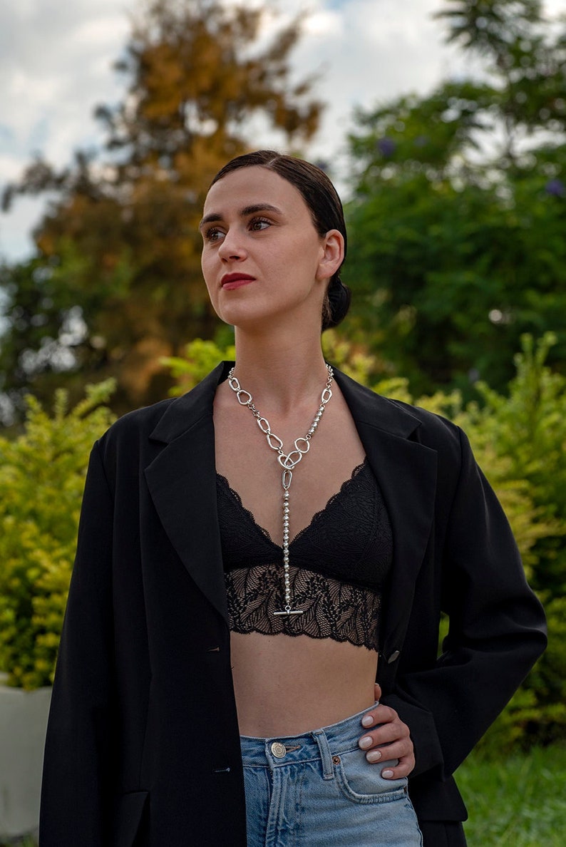 May include: A woman wearing a black blazer, a black lace bralette, and blue jeans. She is wearing a silver chain necklace with a pendant.