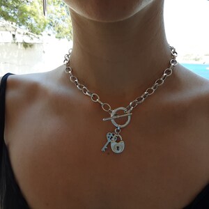 May include: A silver chain necklace with a toggle clasp and a charm featuring a padlock and key.