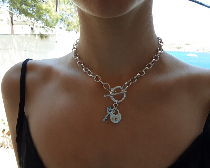 May include: A silver chain necklace with a toggle clasp and a charm featuring a padlock and key.