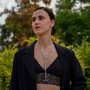 May include: A woman wearing a black blazer and a black lace top with a silver chain necklace. The necklace has a long pendant with a silver chain and a silver toggle clasp.