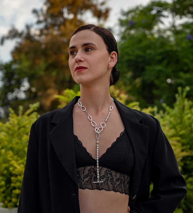 May include: A woman wearing a black blazer and a black lace top with a silver chain necklace. The necklace has a long pendant with a silver chain and a silver toggle clasp.