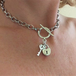 May include: A silver chain necklace with a toggle clasp and a heart-shaped key and padlock charm.
