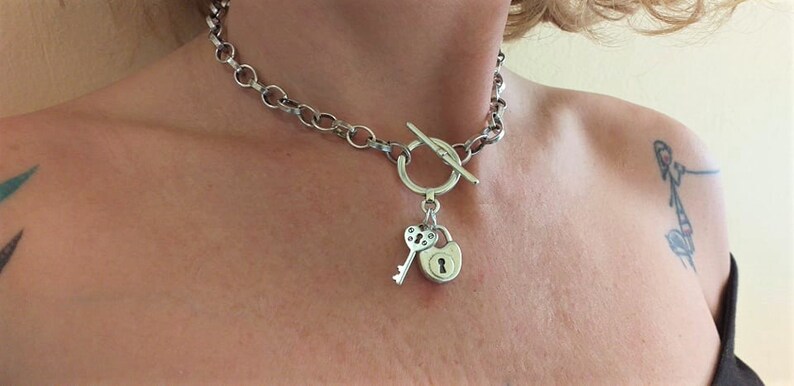 May include: A silver chain necklace with a toggle clasp and a heart-shaped key and padlock charm.